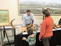 An adult and a child look at a fossil bison skull on a table. Behind the table is a paleontologist talking to guests.