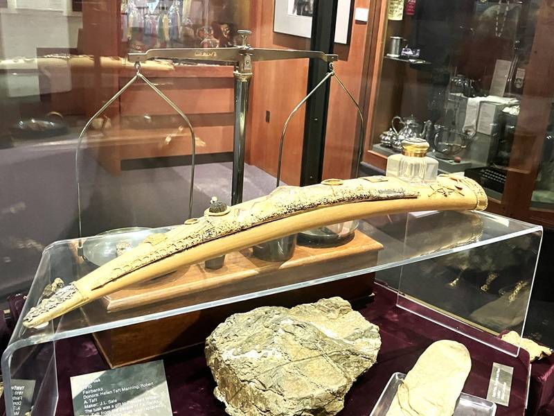 Piece of wood carved in the shape of a walrus tusk, covered in small gold figurines. The piece sits in a museum display case.