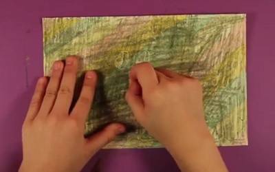Piece of paper covered in black crayon. A hand is using a paperclip to scratch away the black, revealing rainbow colors underneath