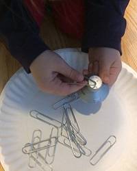 Child's hands holding a magnet over a several paper clips on a paper plate.