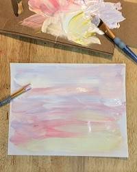 Pastel colored paints on a piece of cardboard, next to a piece of paper painted to look like a winter sunrise.