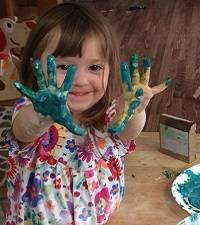 Child smiling at the camera and holding up paint-covered hands.