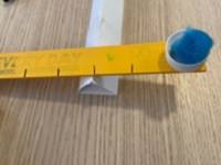 Yellow stick rested on a fulcrum made from a folded piece of paper. A blue pom-pom rests on one end of the stick. The stick has black lines marking one inch intervals.
