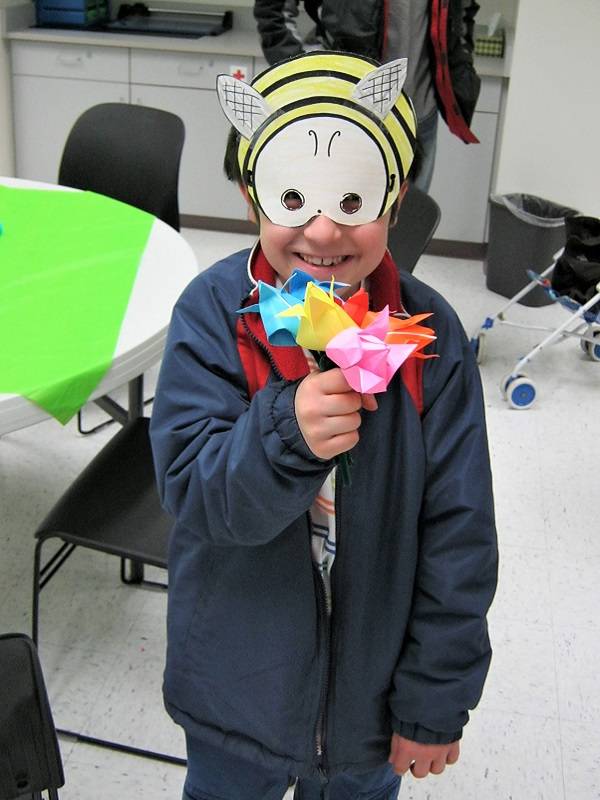Child wearing a paper bumblebee mask and holding origami flowers.