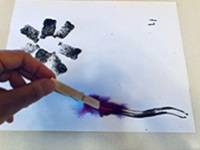 Hand using a homemade paintbrush with feathers on one end to paint on white paper.