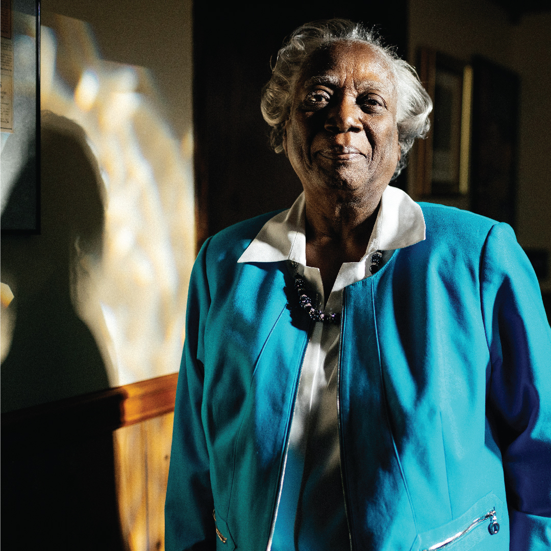 Dorothy Jones stands with the sunlight on her left, casting a shadow. She wears a bright blue blazer and a warm smile. 
