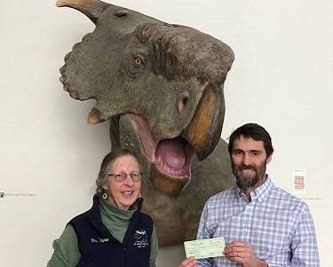 Two people stand in front of a dinosaur sculpture. One is handing a check to the other.