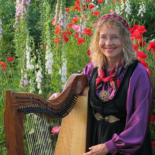 Beth Kolle poses with her harp in Nordic dress in front of red and white flowers