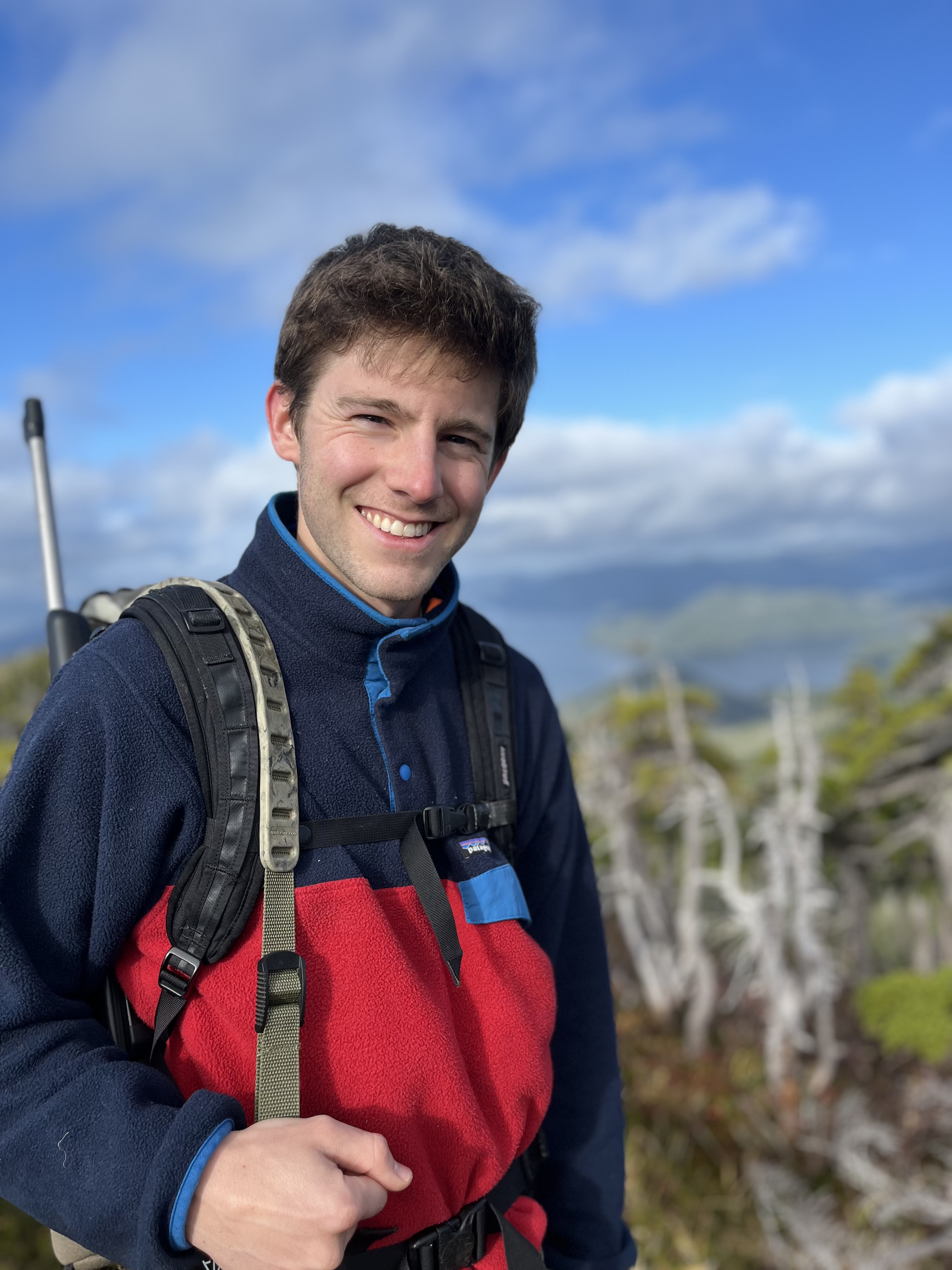 A man in outdoor gear with a rifle over his shoulder smiles in front of a backdrop of mountains and ocean