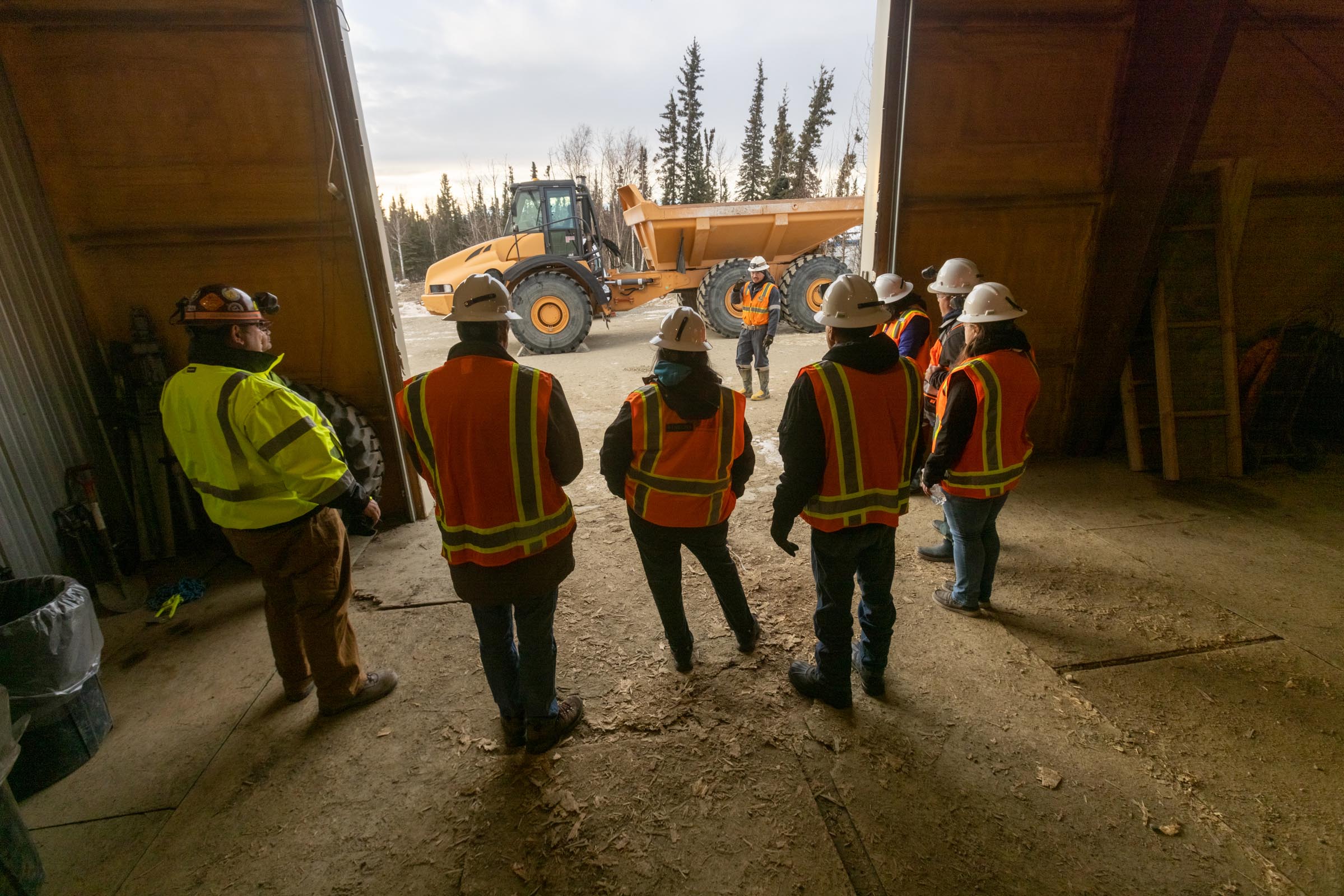 A group of people wearing reflective vests and hard hats listen to a student explain how to safely run heavy equipment.