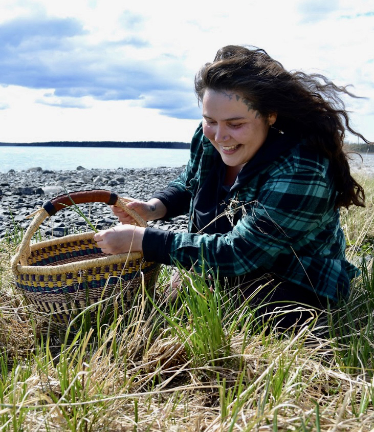 A smiling woman fills a basket with native grasses with a body of water in the background.