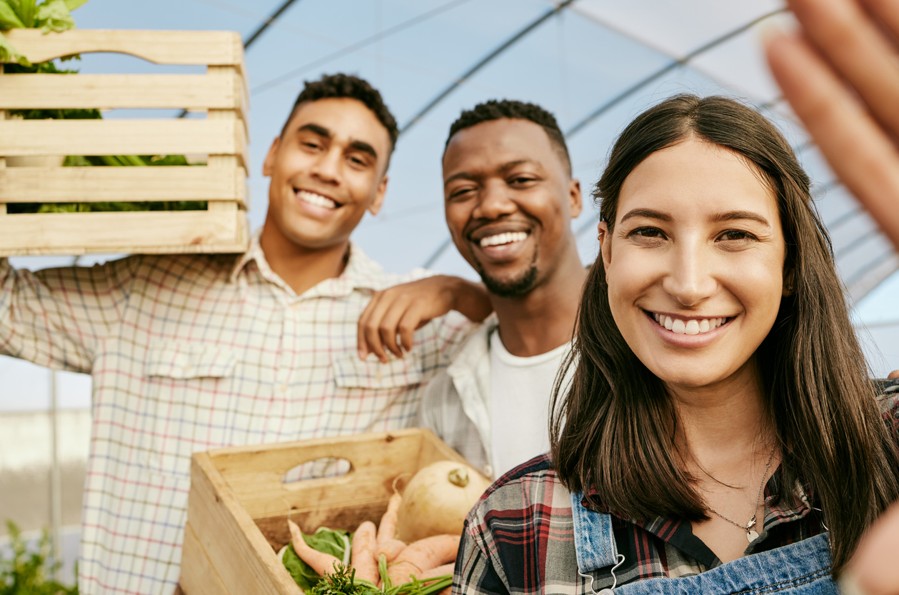 Three smiling young people, two men and a woman, stand in a greenhouse holding crates of fresh vegetables.
