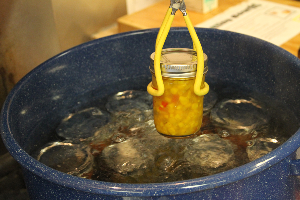 A jar of corn salsa is lowered into a boiling waterbath canner filled with other jars