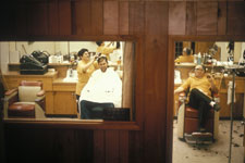A color picture of two men at the barbershop and a woman cutting hair.