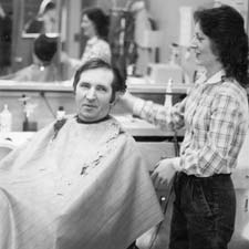 A black and white photograph of a professor having his hair cut by Charlette Lushin.