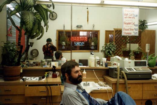 A busy barbershop with a man sitting in the forground with a person taking a picture visible in the mirrors.