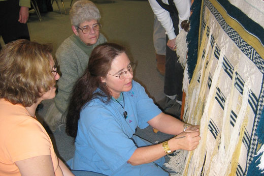 A woman explains some weaving to two other women on an unfinished piece.