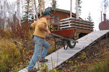 A man pushing a wheelbarrow up a aluminum grate ramp, with a building in the background.