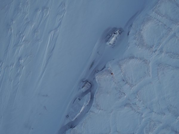Photo courtesy of Ben Jones. UAF researcher Chris Arp, appearing as a dark dot at center right in this aerial photo, stands on Drew Point in late November 2018. Chunks of eroding land lie on the boundary between, at left, snow drifts on the frozen surface of Smith Bay and, to the right, tundra with its characteristic polygon pattern.