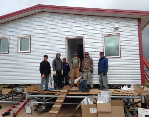 Photo by Molly Rettig. Aaron Cooke, in the T-shirt and blue hat, poses in front of an efficient house he helped design after he and Newtok residents built it at the new village site of Mertarvik in 2016.