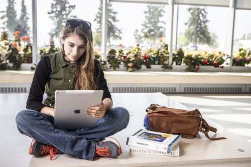 young woman working on a tablet while sitting cross-legged by windows