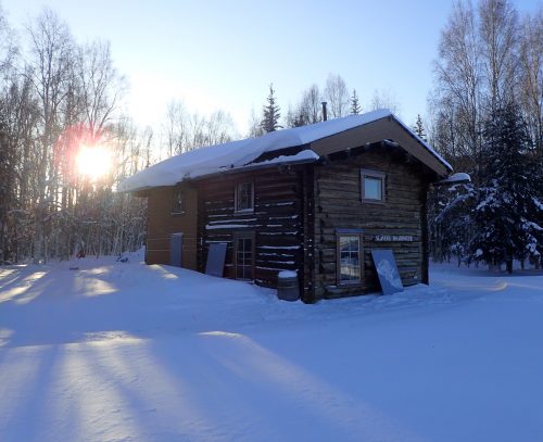 Photo by Bob Gillis. Sunlight dapples the snow by Slaven’s Roadhouse, a restored 1932 structure on the Yukon River used by National Park Service rangers as a hospitality stop during the Yukon Quest sled dog race.