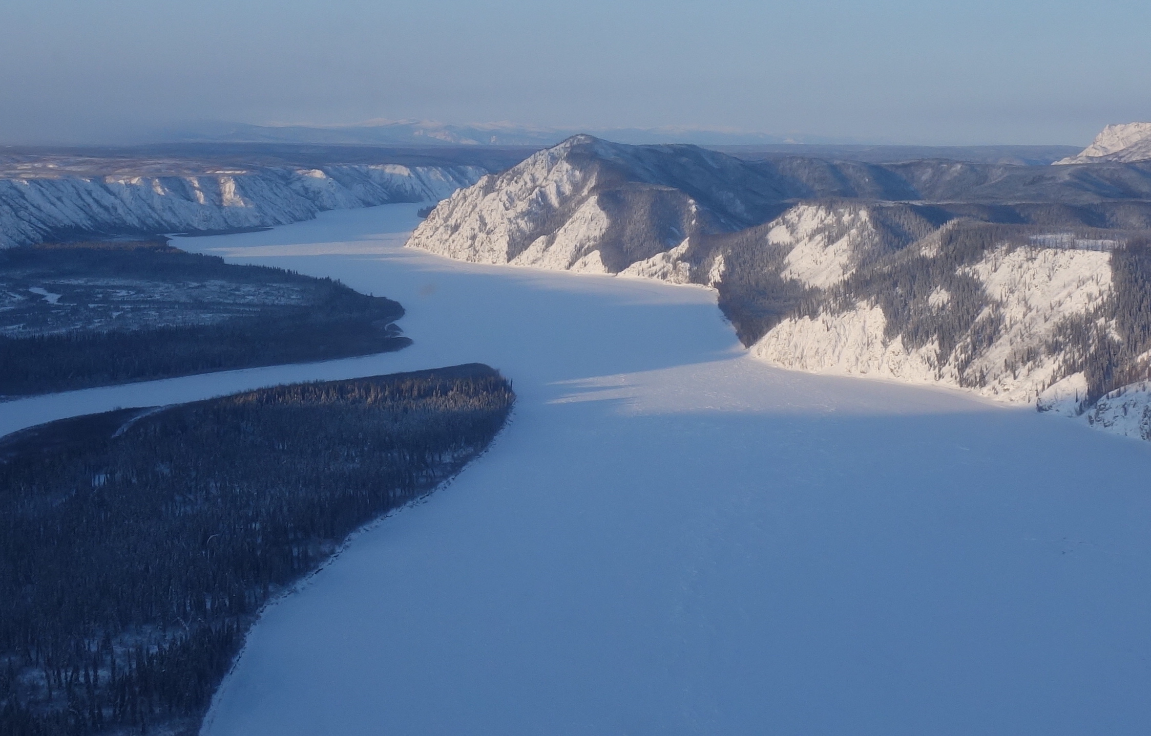 Photo by Adam Bucki. The Yukon River, pictured here in early February 2020, flows westward just downstream from Slaven’s Roadhouse. Woodchopper Bluff is to the right.