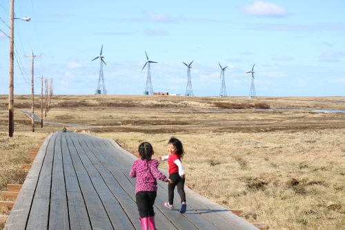 Photo by Amanda Byrd. Children run along a boardwalk in Kongiganak with wind turbines in the background.