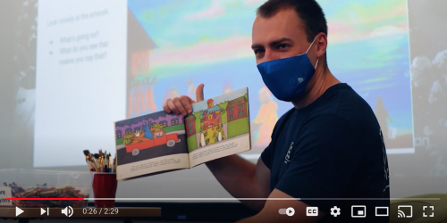 Video screen shot of a man holding up a children's book. He is wearing a mask..