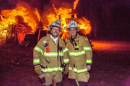 Two men in firefighting gear stand in front of massive bonfires in an empty field.