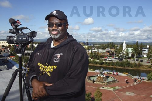 Cover image of a man standing beside a TV camera overlooking downtown Fairbanks in summer