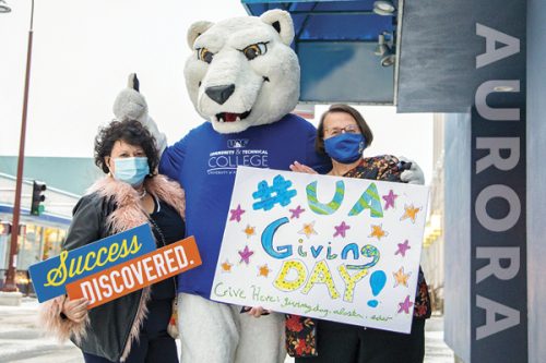 Two masked students with signs celebrating UAF and UAF giving day stand with a T-shirt-clad Nanook mascot.