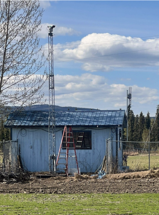 A piece of equipment on a long pole sits above a light blue farm outbuilding next to a tree.