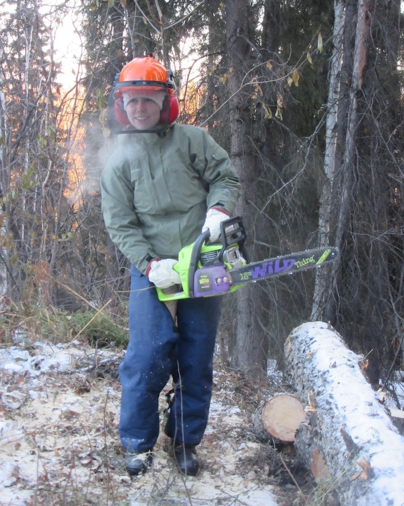 A woman wearing safety gear, including gloves, a helmet, face mask and ear protection, cuts a log with a chainsaw.