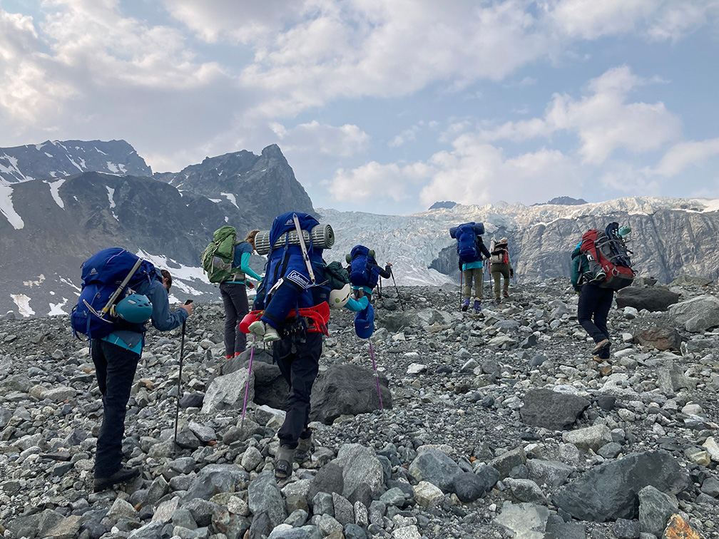 A group of young women facing away from the camera and wearing heavy packs ascend a scree-covered slope at the base of a glacier.