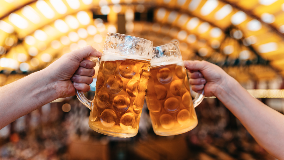 Two hands clinking glasses in front of the Oktoberfest marquee.