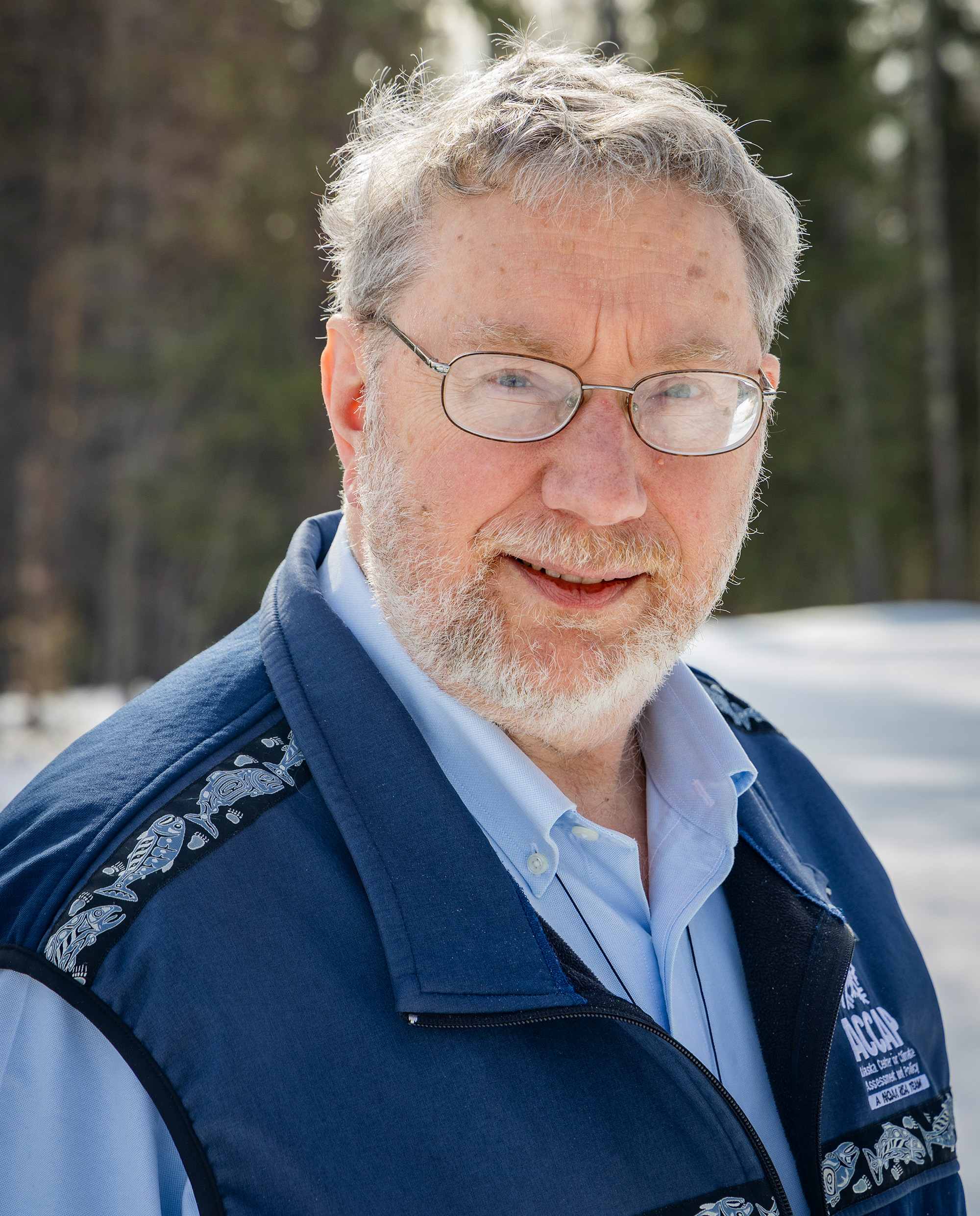 A man with graying hair wearing glasses and a button up shirt and fleece vest outdoors in the snow.