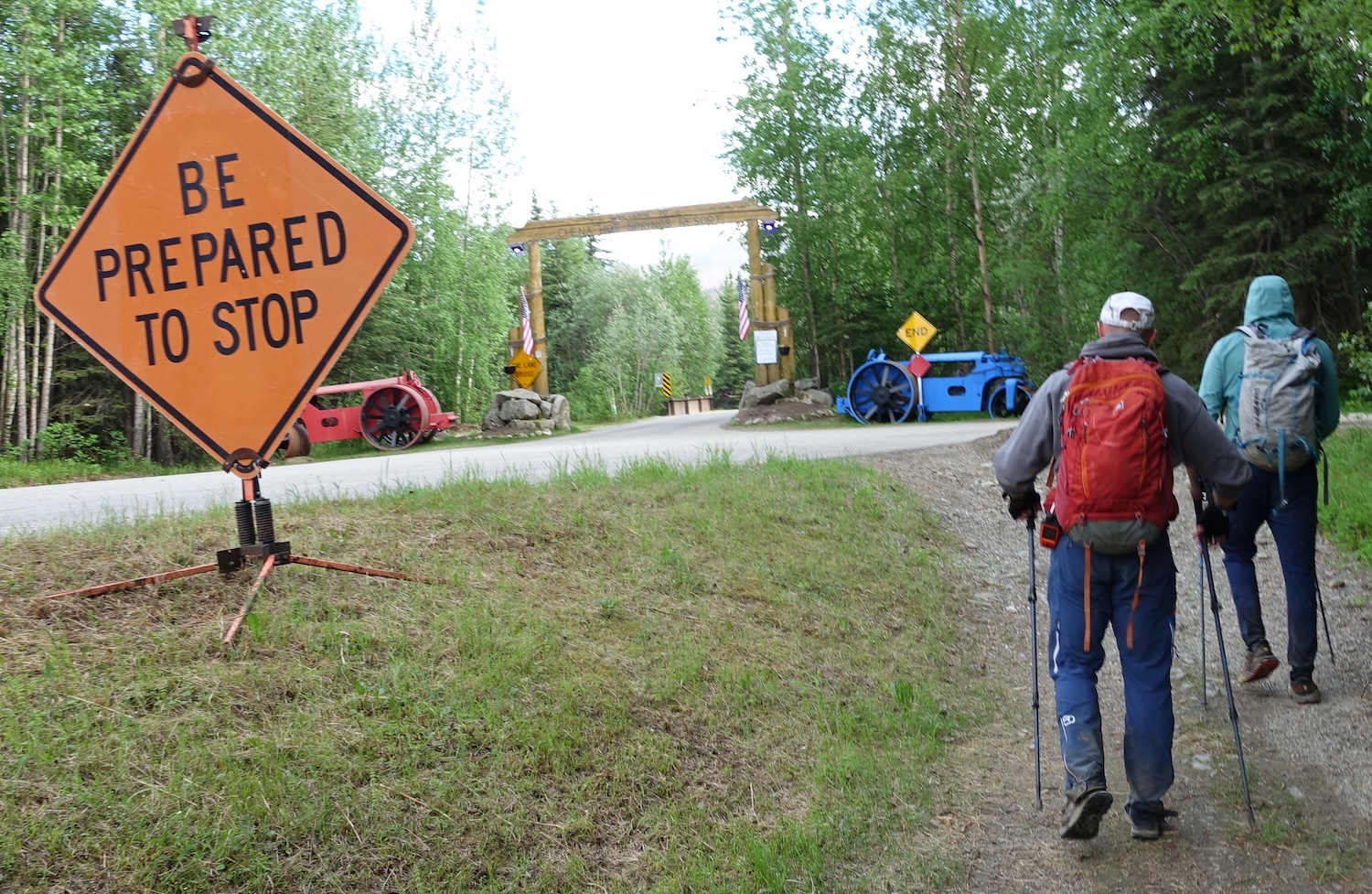 Two men walk by an orange, diamond-shaped highway sign that reads "Be prepared to stop." A log arch with the words "Chena Hot Springs Resort" on it rises above a road in the background. Old tractors, one red and one blue, sit on either side of the arch.