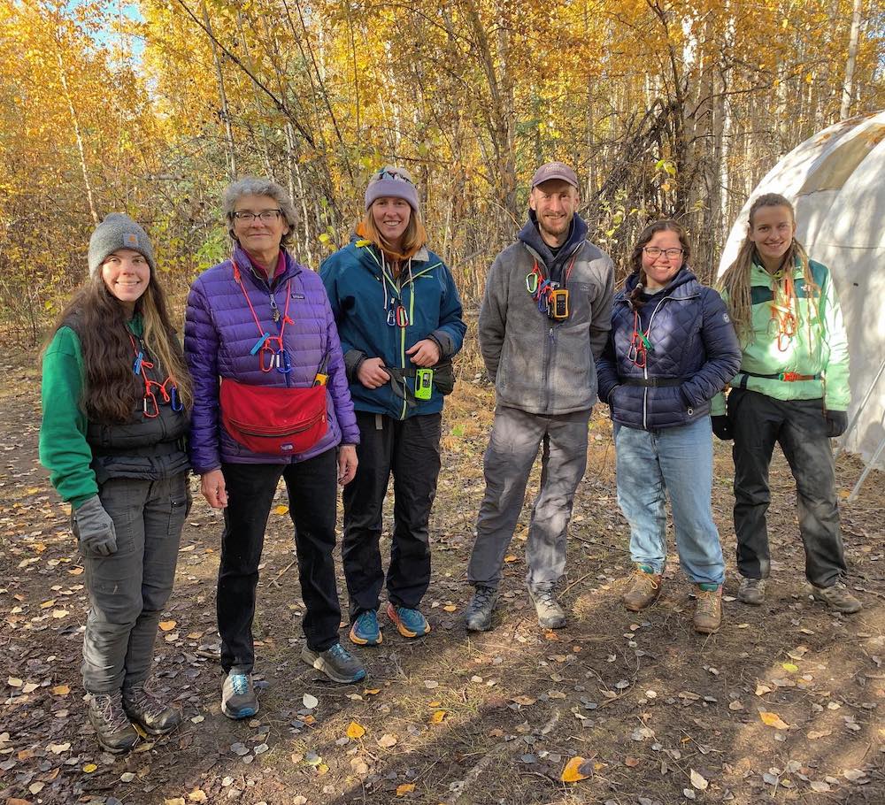 Five women and a man, all of varying ages and dressed in coats, stand together in a forest of deciduous trees in yellow fall foliage. Hanging from their necks are strings, each with two carabiner clips and a clothespin attached. A frame tent stands behind them on the right.