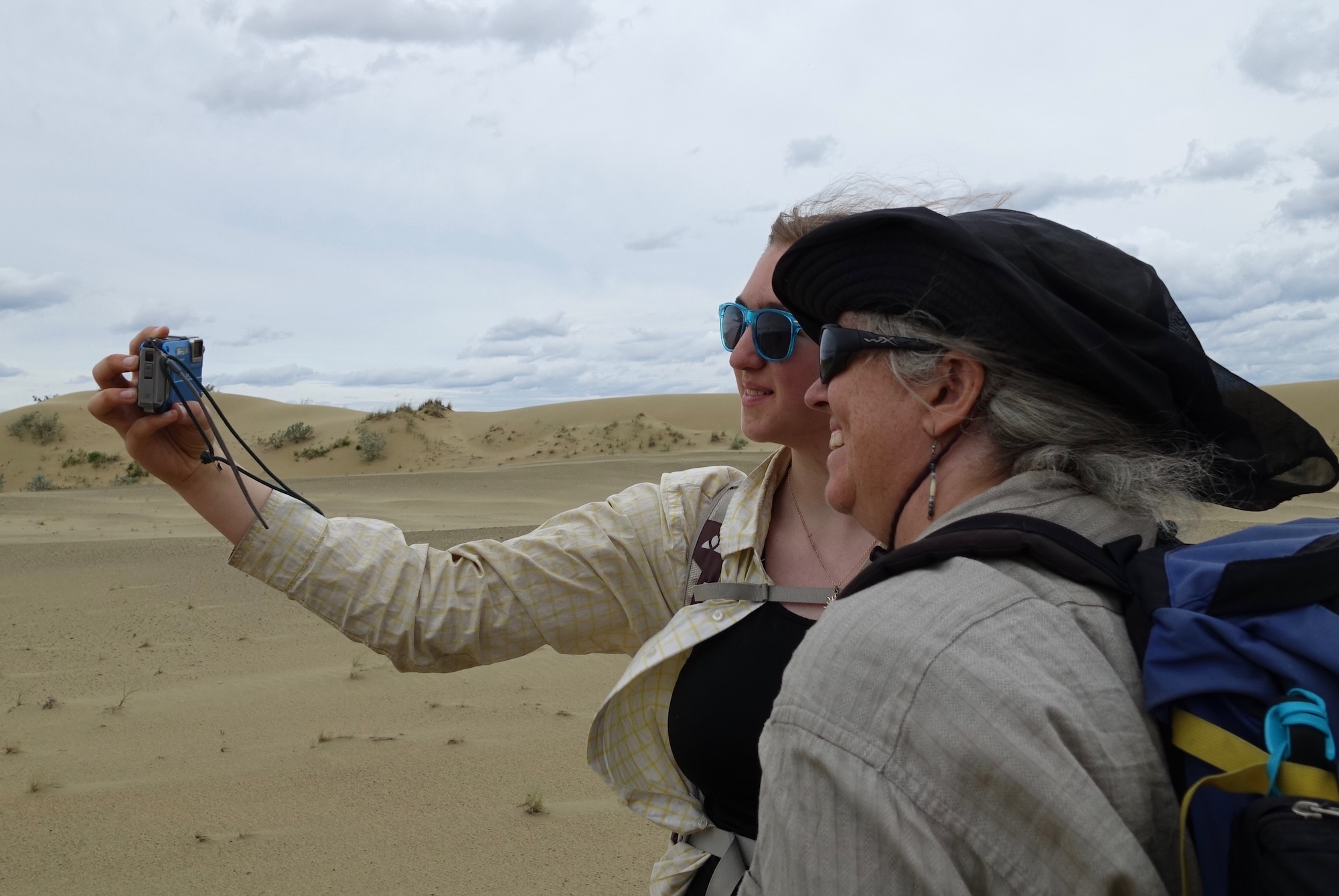 Two women smile while taking a self-portrait in an expanse of sand studded with short vegetation.
