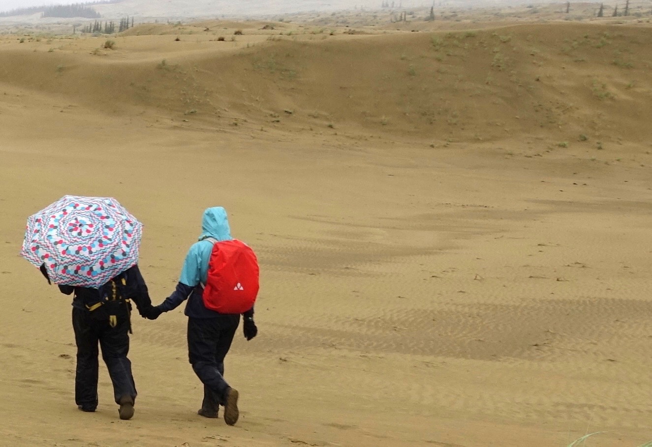 Two women, one carrying an umbrella, walk on sand dunes.