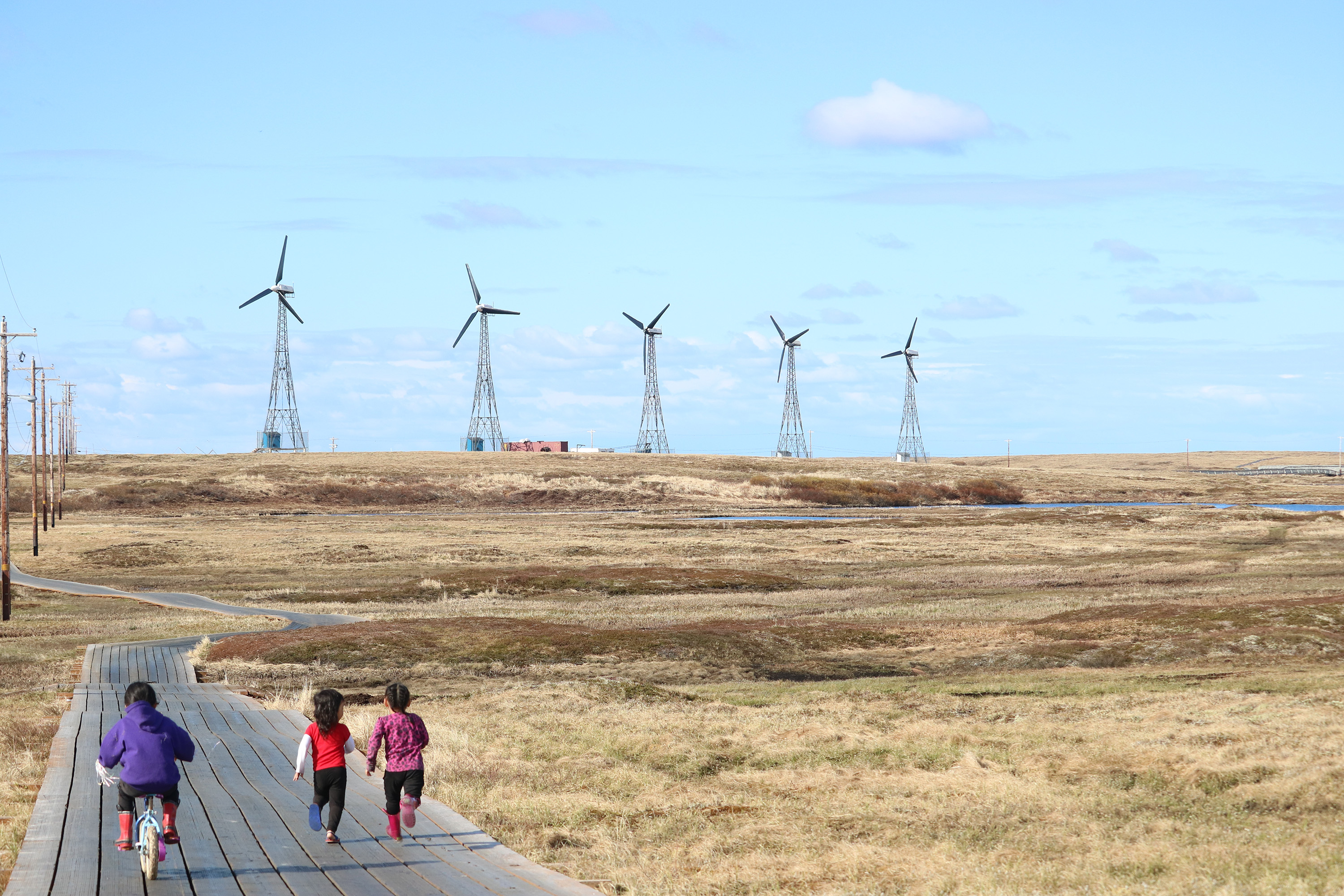 Children run along a boardwalk in the village of Kongiganak with the community’s 475 kw wind farm in the distance
