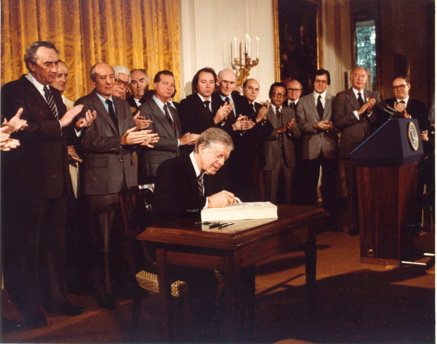 A man in a suit sits at a wooden desk signing a document while surrounded by other men in suits applauding.