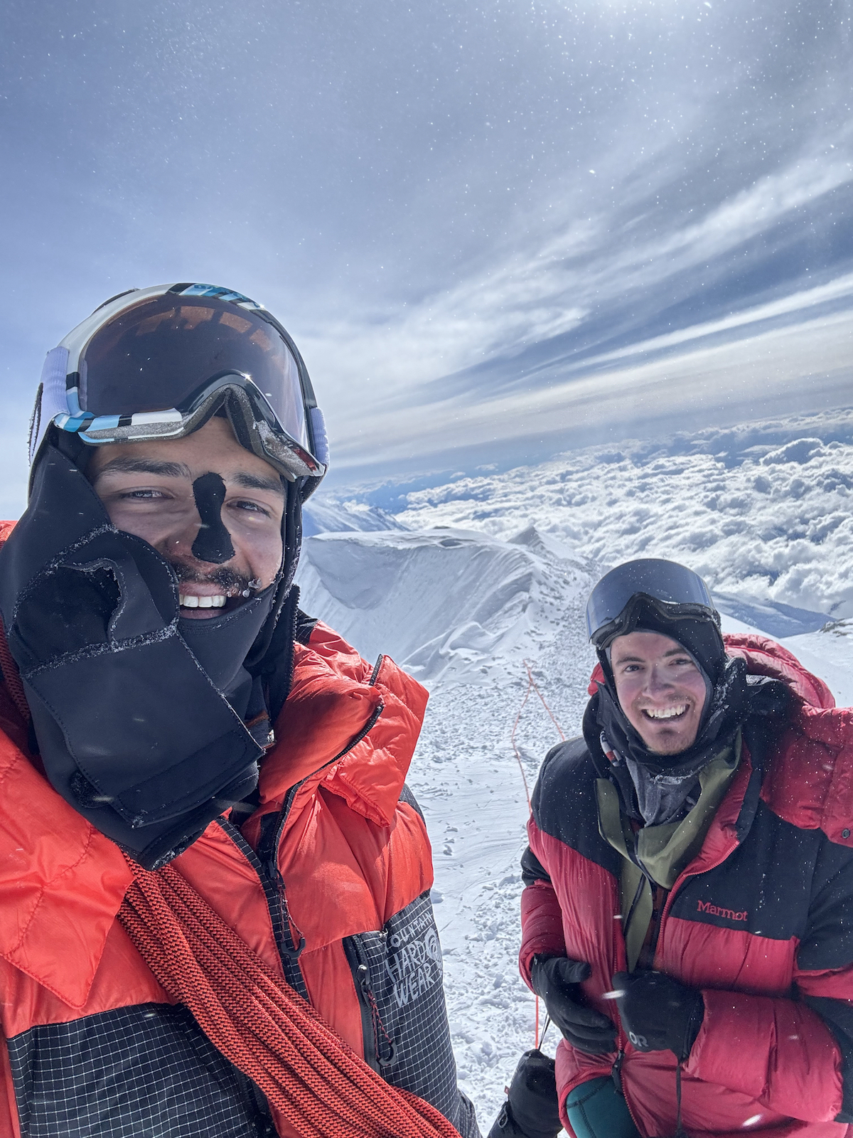 Two men in winter gear stand on a high mountain peak.