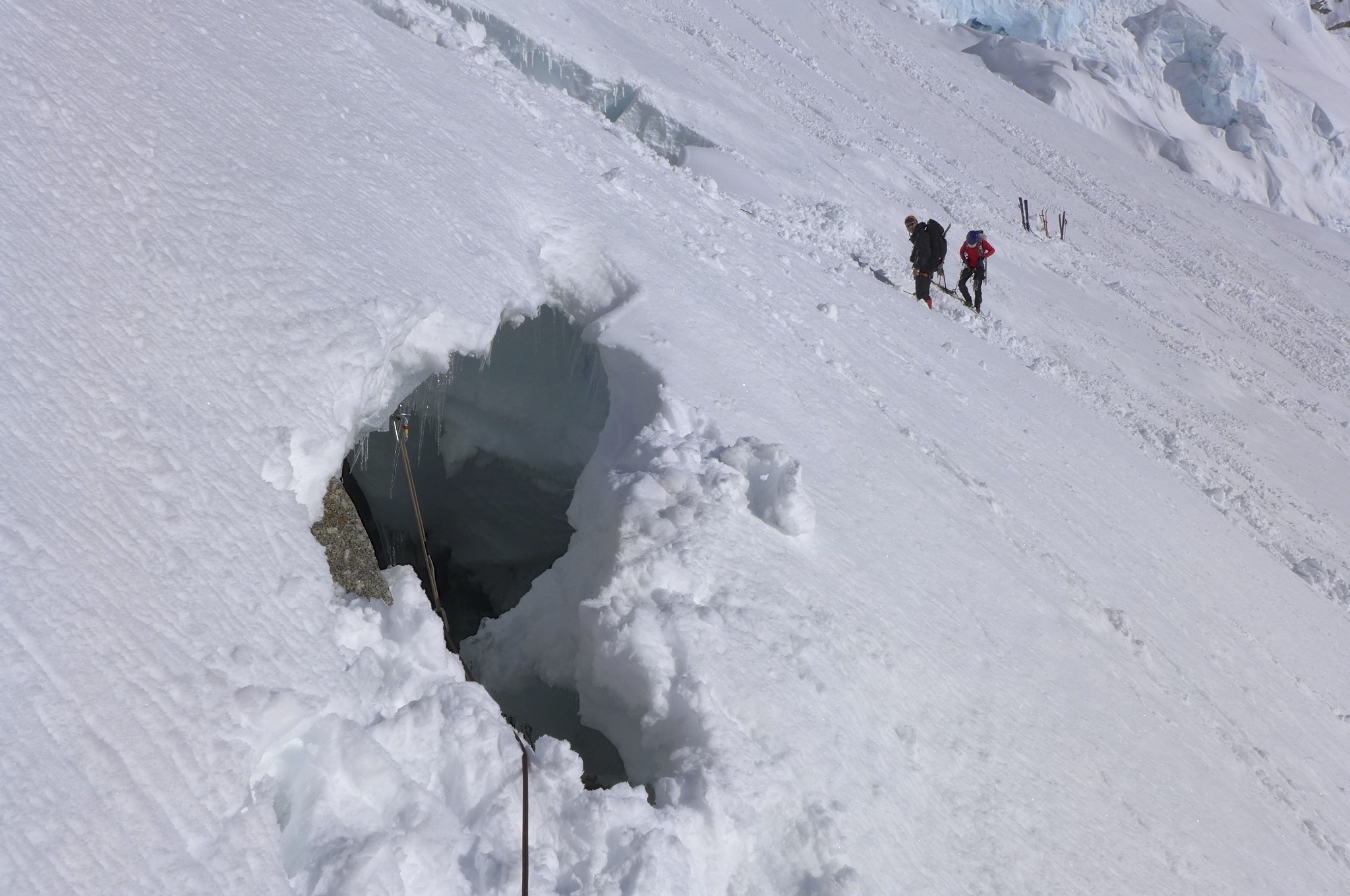 A hole drops into a steeply sloped, snow-covered glacier. Two people stand in the background on the glacier.