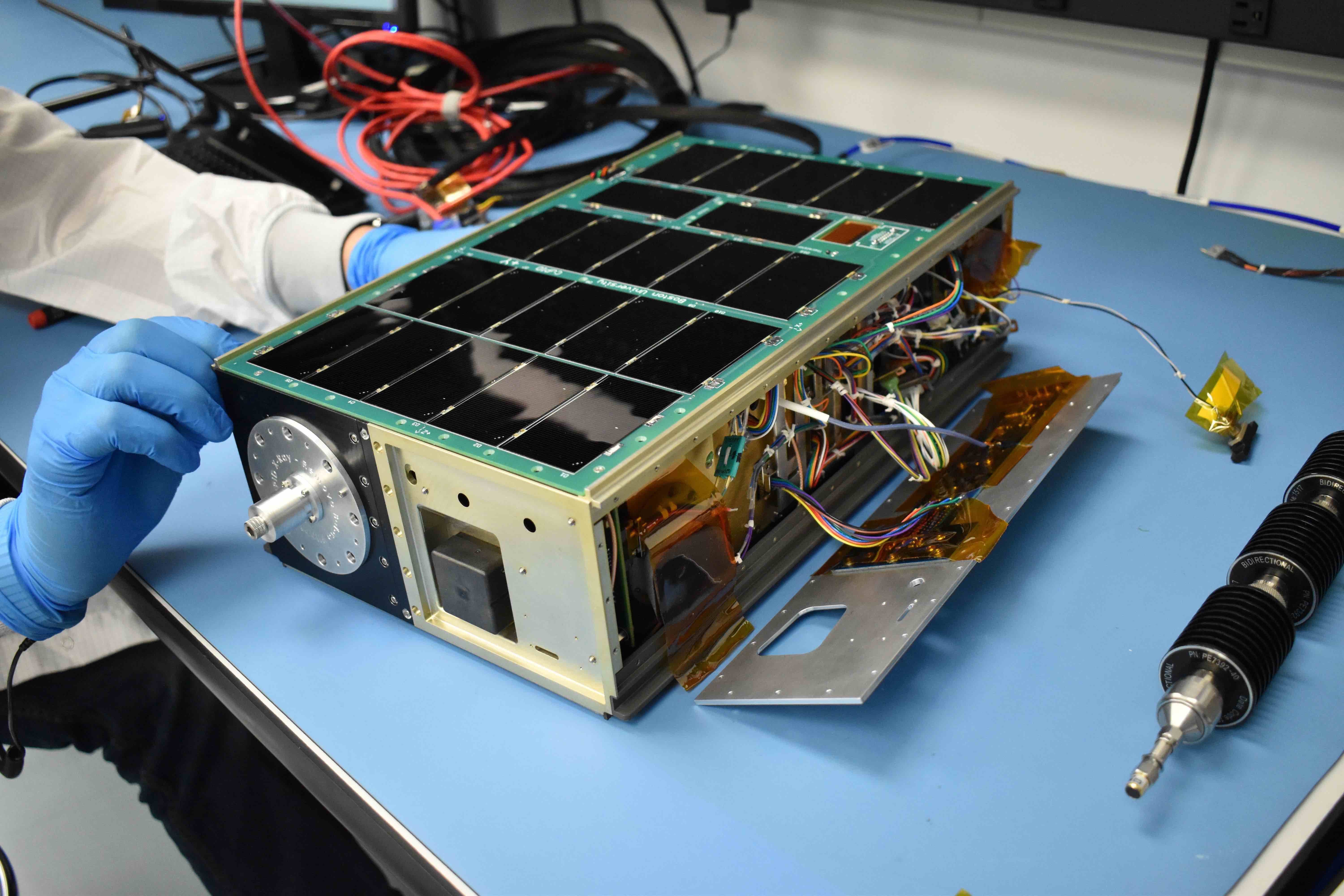 A person in gloves works on a small satellite, called a cubesat, on a table.