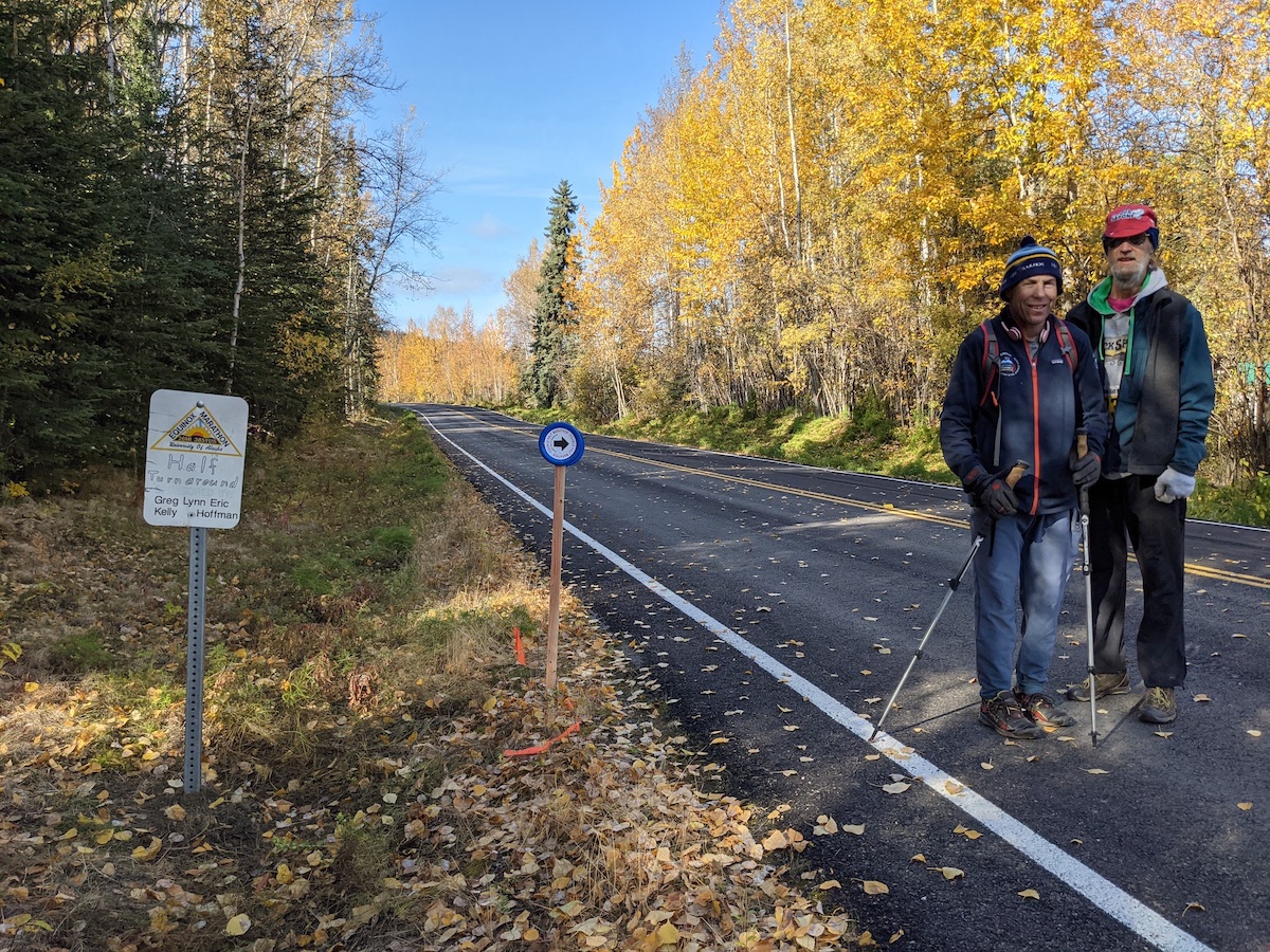 In the right side of the photo, two men stand on a paved road with golden trees in the background and golden leaves on the ground and pavement. At left, an Equinox Marathon sign marks the halfway turnaround point and an blue circular sign on a wooden stake sports an arrow pointing to the right.