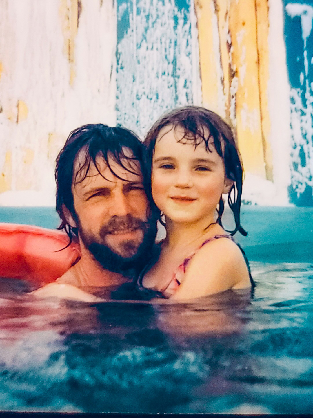 A bearded man holds a young girl in a swimming pool.
