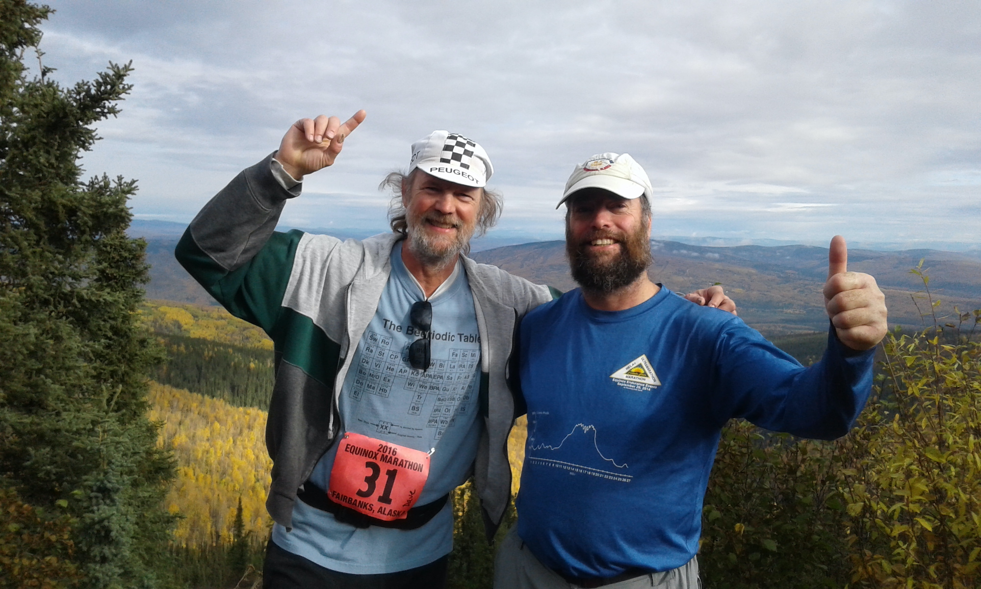 On a high hillside, two bearded men in white hats stand with their arms around each others' shoulders and point to the sky. Behind them is a view of rolling hills covered with golden fall vegetation. 