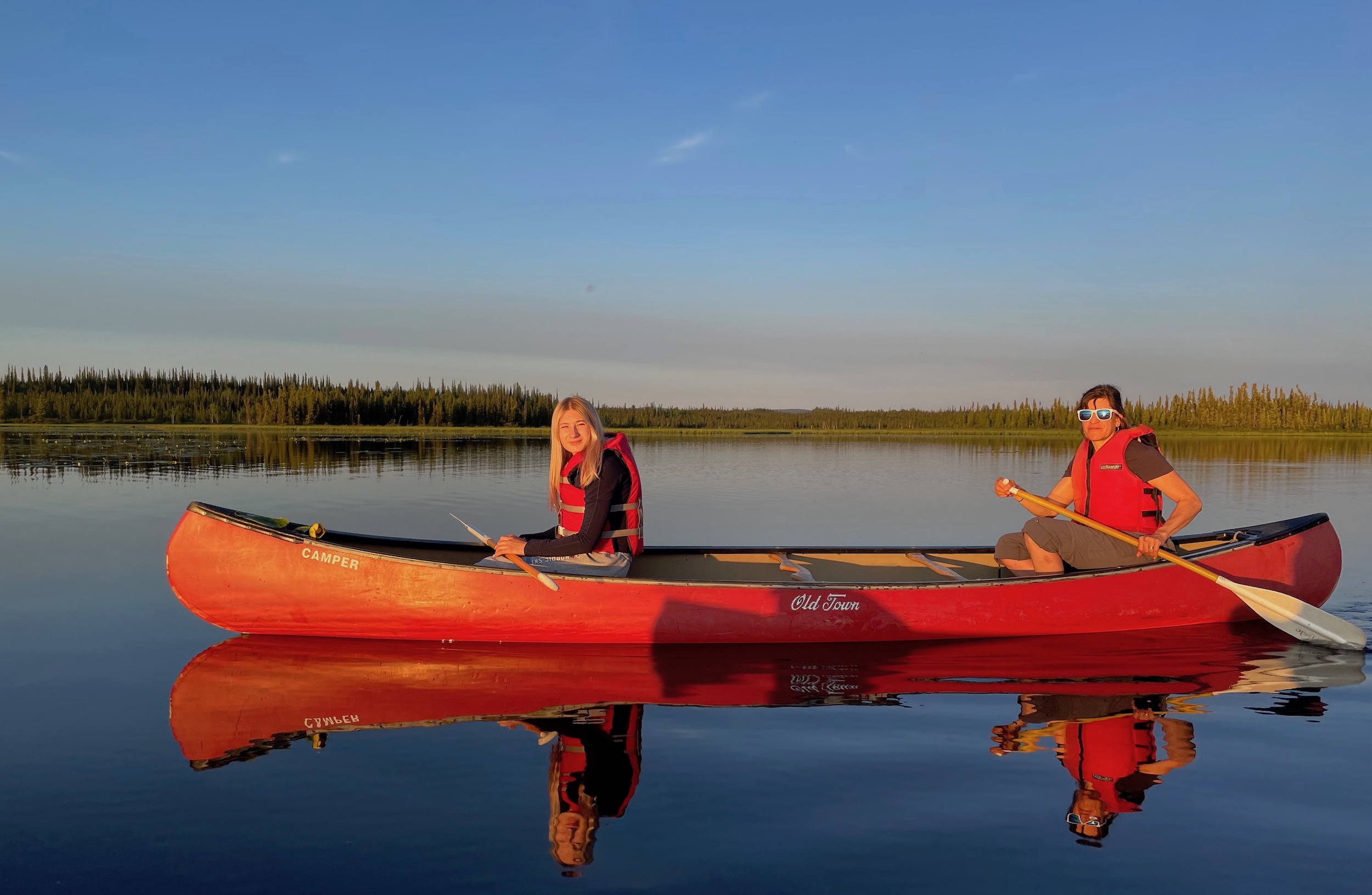 In orange sunshine, two women in life jackets paddle a red canoe across a smooth lake with a line of spruce on a far shoreline in the background.